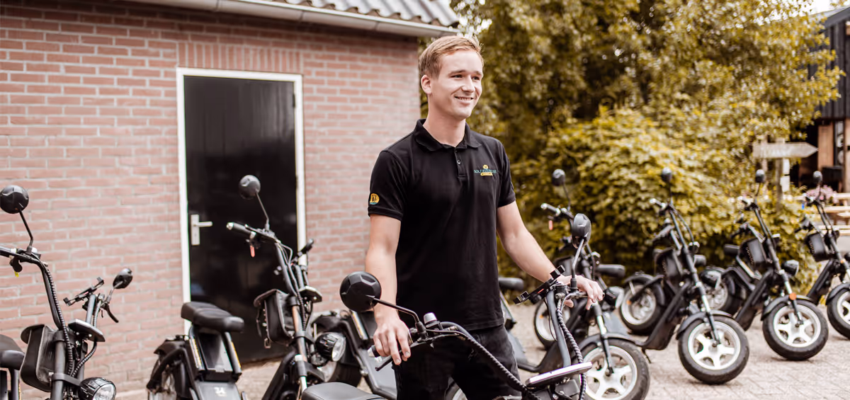 Een vriendelijke medewerker staat lachend tussen een rij e-choppers voor een rode bakstenen schuur, klaar om gasten te verwelkomen voor een rit.