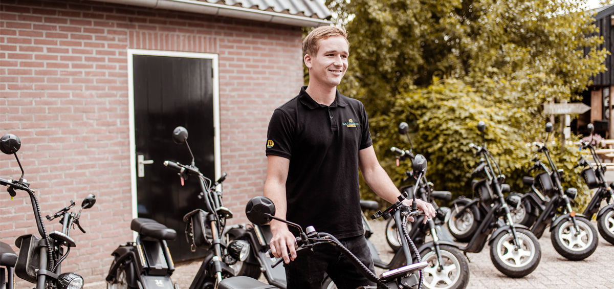 Een vriendelijke medewerker staat lachend tussen een rij e-choppers voor een rode bakstenen schuur, klaar om gasten te verwelkomen voor een rit.