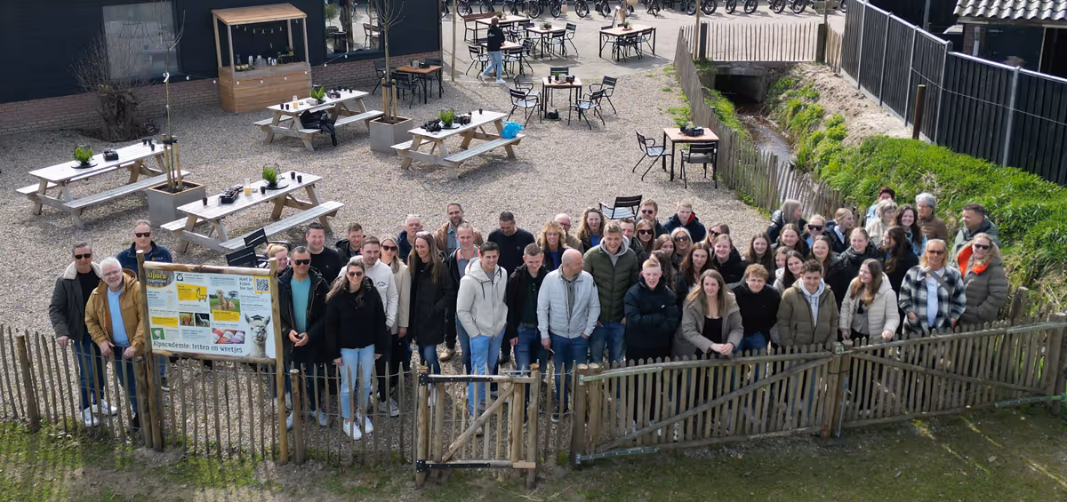 Een grote groep mensen poseert lachend voor een houten hek op een gezellig erf met picknicktafels en een klein stroompje, klaar voor een gezamenlijk uitje of activiteit.