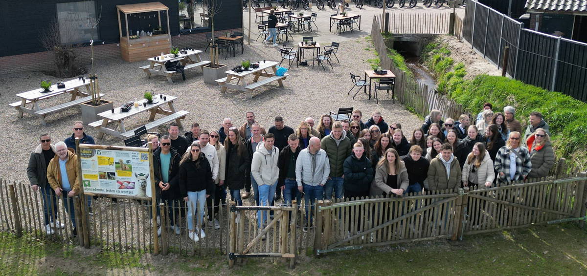 Een grote groep mensen poseert lachend voor een houten hek op een gezellig erf met picknicktafels en een klein stroompje, klaar voor een gezamenlijk uitje of activiteit.
