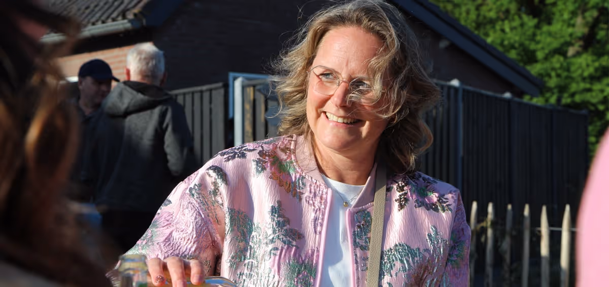 Een vrouw met een glimlach serveert iets in de buitenlucht, gekleed in een opvallend roze jasje met bloemenpatroon, terwijl de zon op haar gezicht schijnt en mensen op de achtergrond gezellig samen zijn.