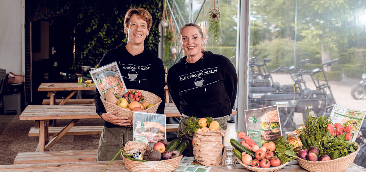 Twee lachende vrouwen in zwarte “buitenkookfeest.nl”-truien presenteren manden met verse groenten en fruit op een houten tafel in een sfeervolle tent met e-choppers op de achtergrond.