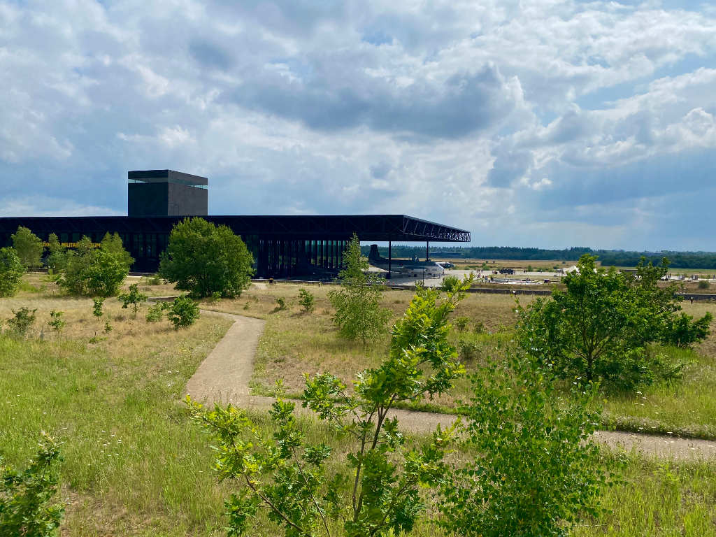 Modern zwart hangargebouw in een weids grasveld met een militair transportvliegtuig onder de overkapping, een slingerend wandelpad in de voorgrond en wolkenlucht erboven.
