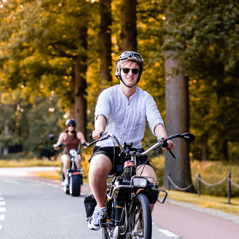 Jonge man met zonnebril en helm rijdt op een brede elektrische chopper over een landelijk fietspad, met een vrouw op een e-chopper op de achtergrond en bomen in de avondzon.
