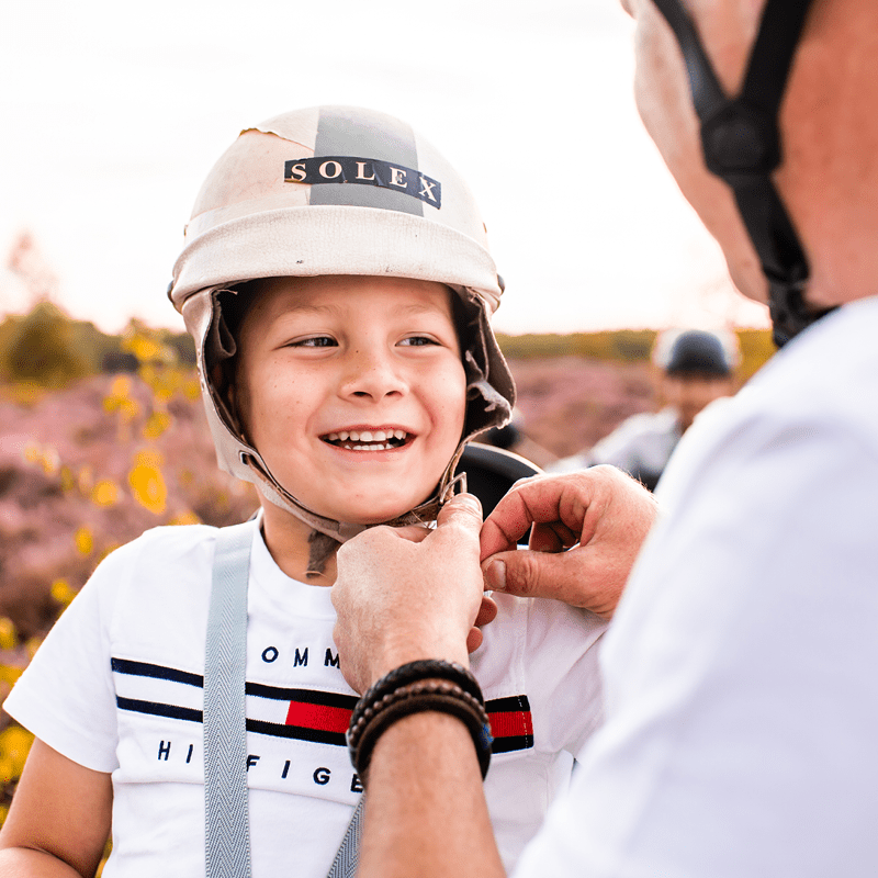 Blakende jongen met een witte “SOLEX” helm lacht terwijl een volwassene zijn kinbandje vastmaakt voor een rit door de paarse hei.