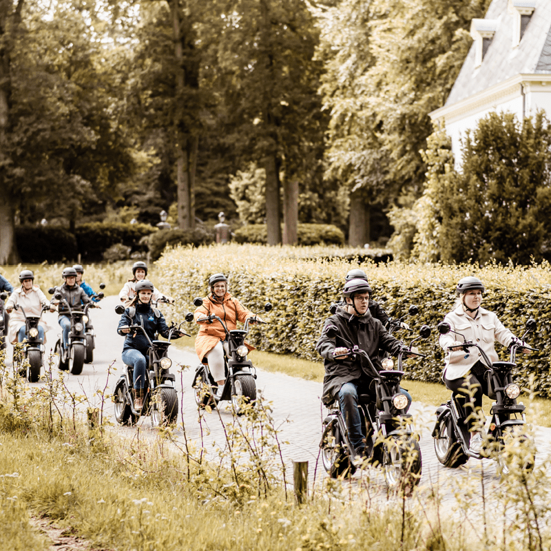 Een groep mensen rijdt in vrolijke stoet op e-choppers langs een statig landhuis en groene hagen op een zonnig landweggetje.