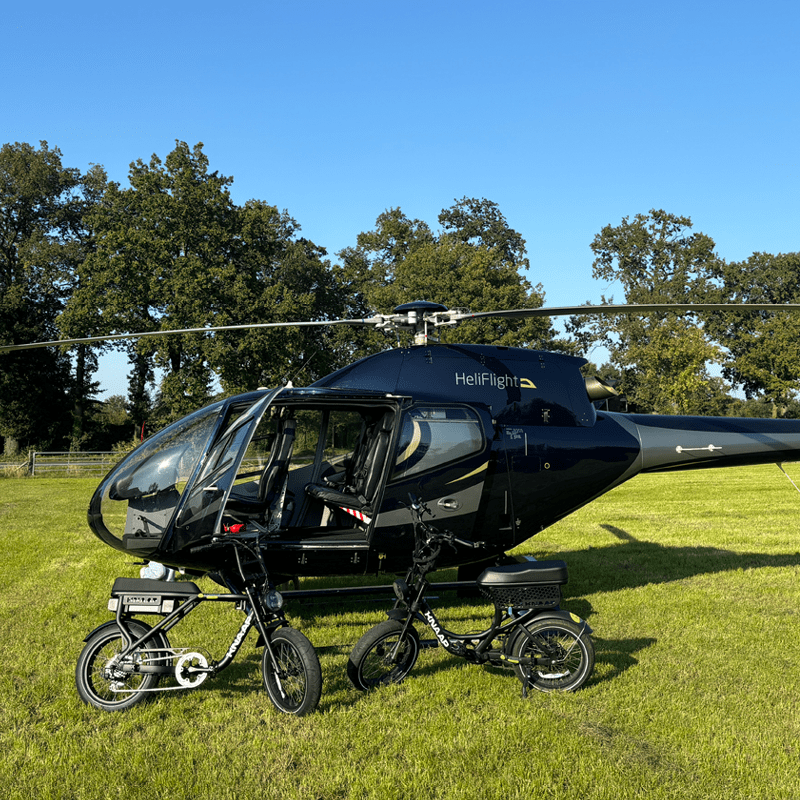 Zwarte helikopter met open deur staat op een grasveld, met twee elektrische fietsen ervoor en bomen op de achtergrond onder een heldere blauwe lucht.