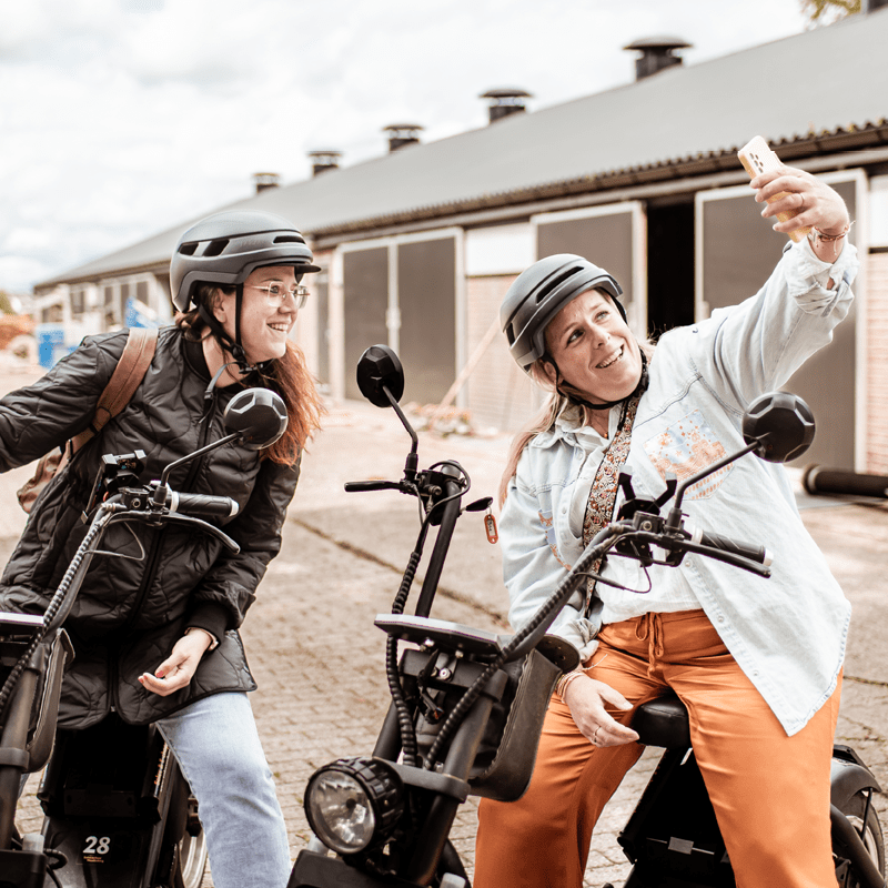 Twee vrouwen met helmen zitten op elektrische choppers, lachtend terwijl de rechter een selfie maakt voor een rij stallen.