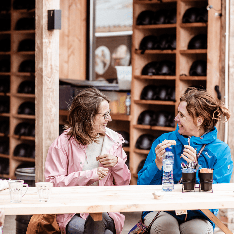 Twee vrouwen in kleurrijke jassen zitten lachend aan een houten tafel bij een helmrek, genietend van een snack en een fles water in een buitenatelier-achtige setting.