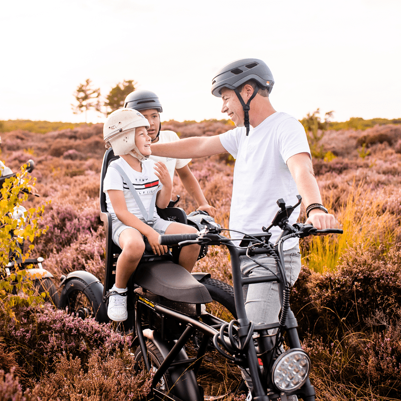 Blije vader in helm praat met zijn zoon op de bagagedrager van een e-chopper, omringd door paarse heidevelden tijdens een zonnige rit.
