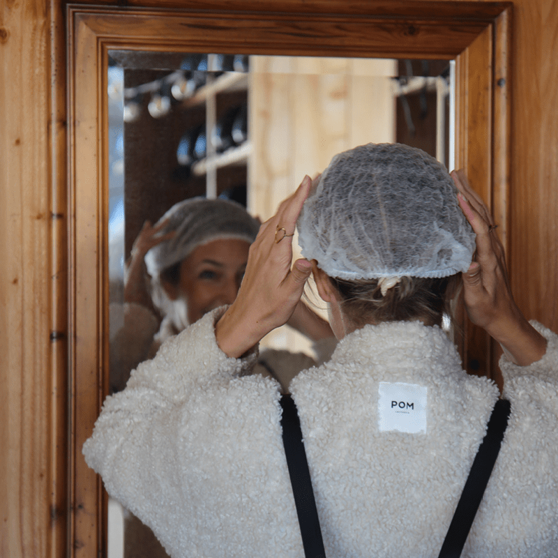 Vrouw van achteren gefotografeerd terwijl ze haar witte wegwerphairtube voor een spiegel rechttrekt in een houten ruimte met helmen op de achtergrond.