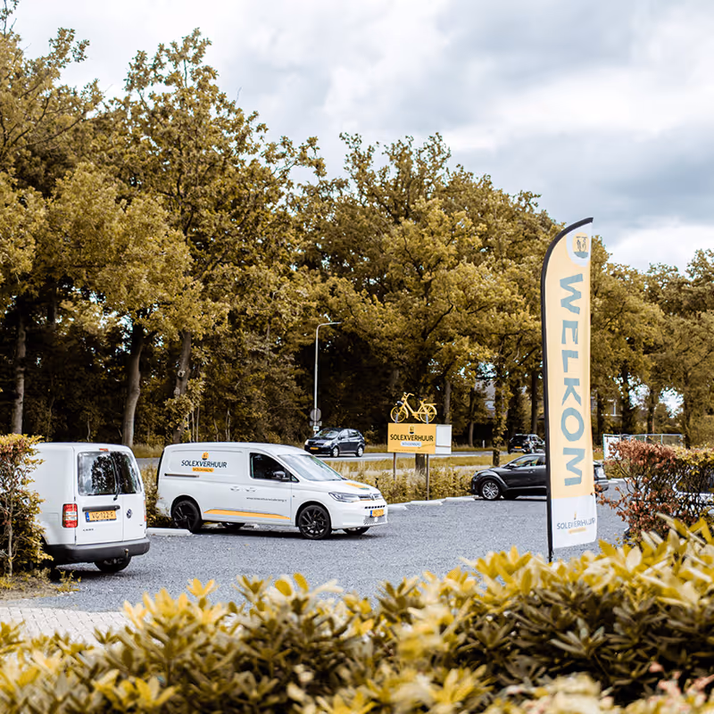 Parkeerterreintje met twee bedrijfsbusjes naast een vlag met “WELKOM” en een bord met daarop een geel fietslogo, gelegen langs een rustige laan met bomen.