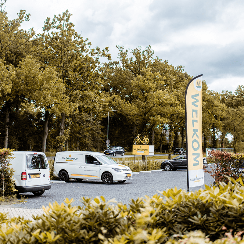 Parkeerterreintje met twee bedrijfsbusjes naast een vlag met “WELKOM” en een bord met daarop een geel fietslogo, gelegen langs een rustige laan met bomen.