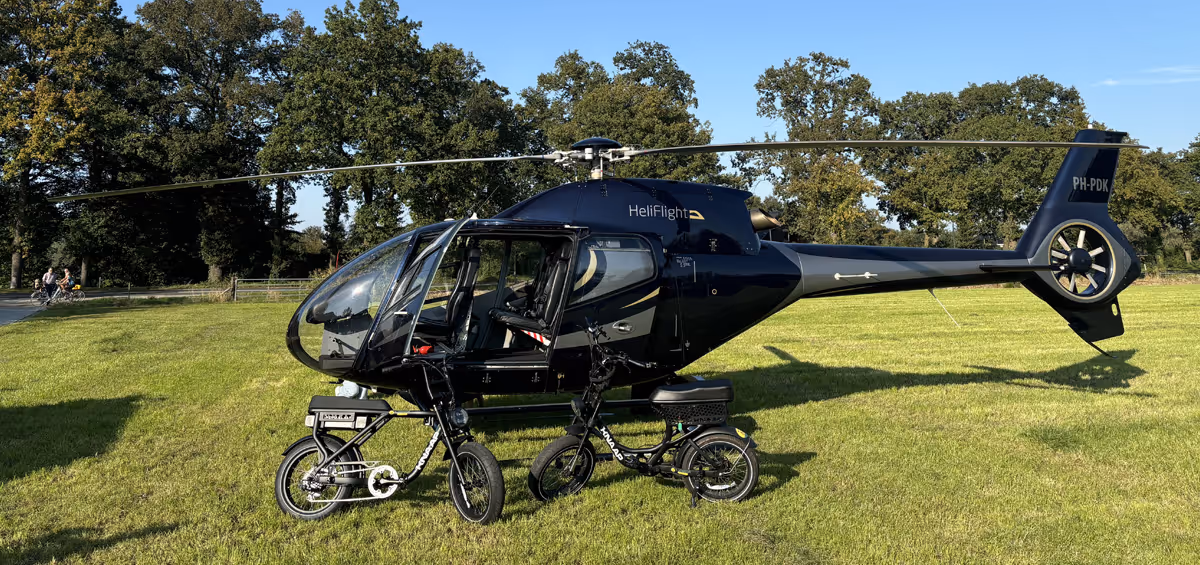 Zwarte helikopter met open deur staat op een grasveld, met twee elektrische fietsen ervoor en bomen op de achtergrond onder een heldere blauwe lucht.