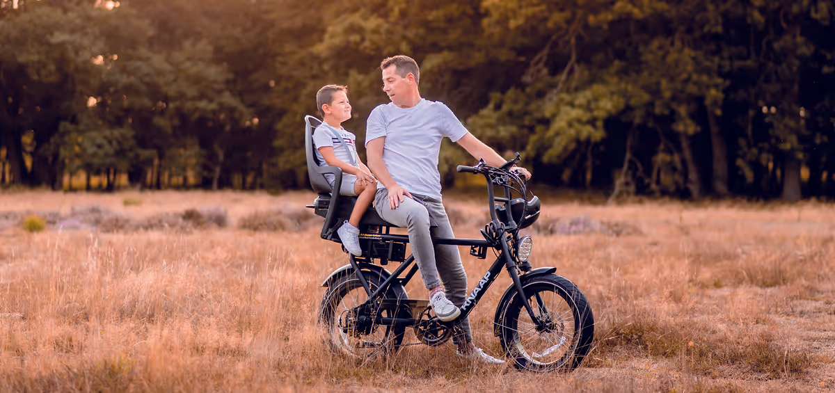 Vader en zoon in helm zitten samen op een elektrische fatbike, met de zoon op een kinderzitje achterop, genietend van een rit door een gouden heideveld bij zonsondergang.