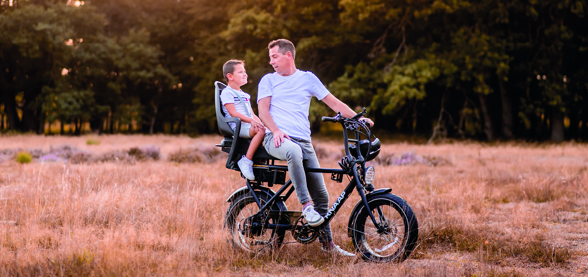 Vader en zoon in helm zitten samen op een elektrische fatbike, met de zoon op een kinderzitje achterop, genietend van een rit door een gouden heideveld bij zonsondergang.