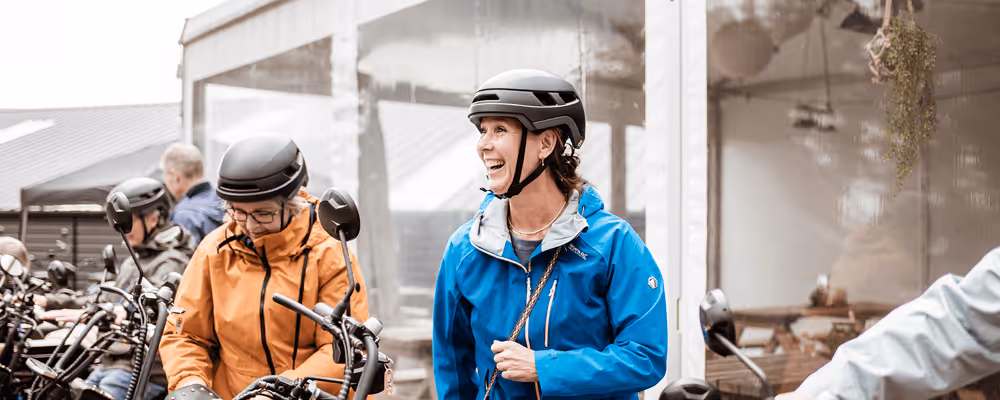 Vrouw in blauwe jas en helm lacht tijdens het opstappen op een e-chopper naast andere mensen met helmen en fietsen voor een tent met picknicktafels.
