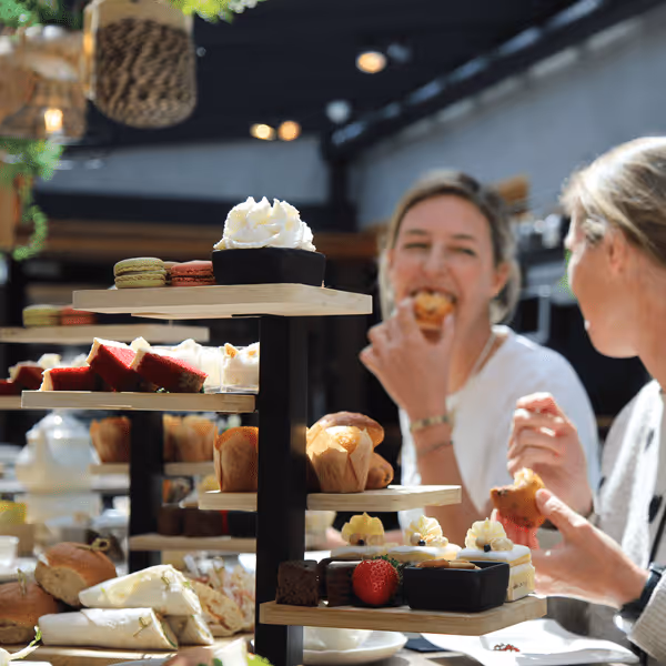 Drieëenheid houten etagères vol hapklare zoetigheden zoals macarons, cakejes, muffins en slagroom, terwijl twee vrouwen erachter genieten van hun afternoon tea in een zonnige, groene setting.