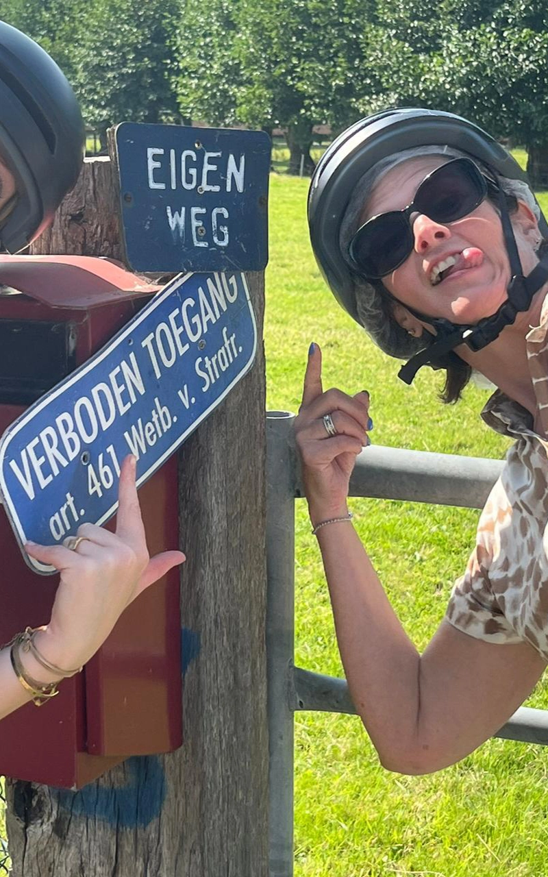 Een vrouw met een helm die wijst naar een blauw bordje met de 'Eigen weg' erop. Ze staan in een weiland bij een hek.