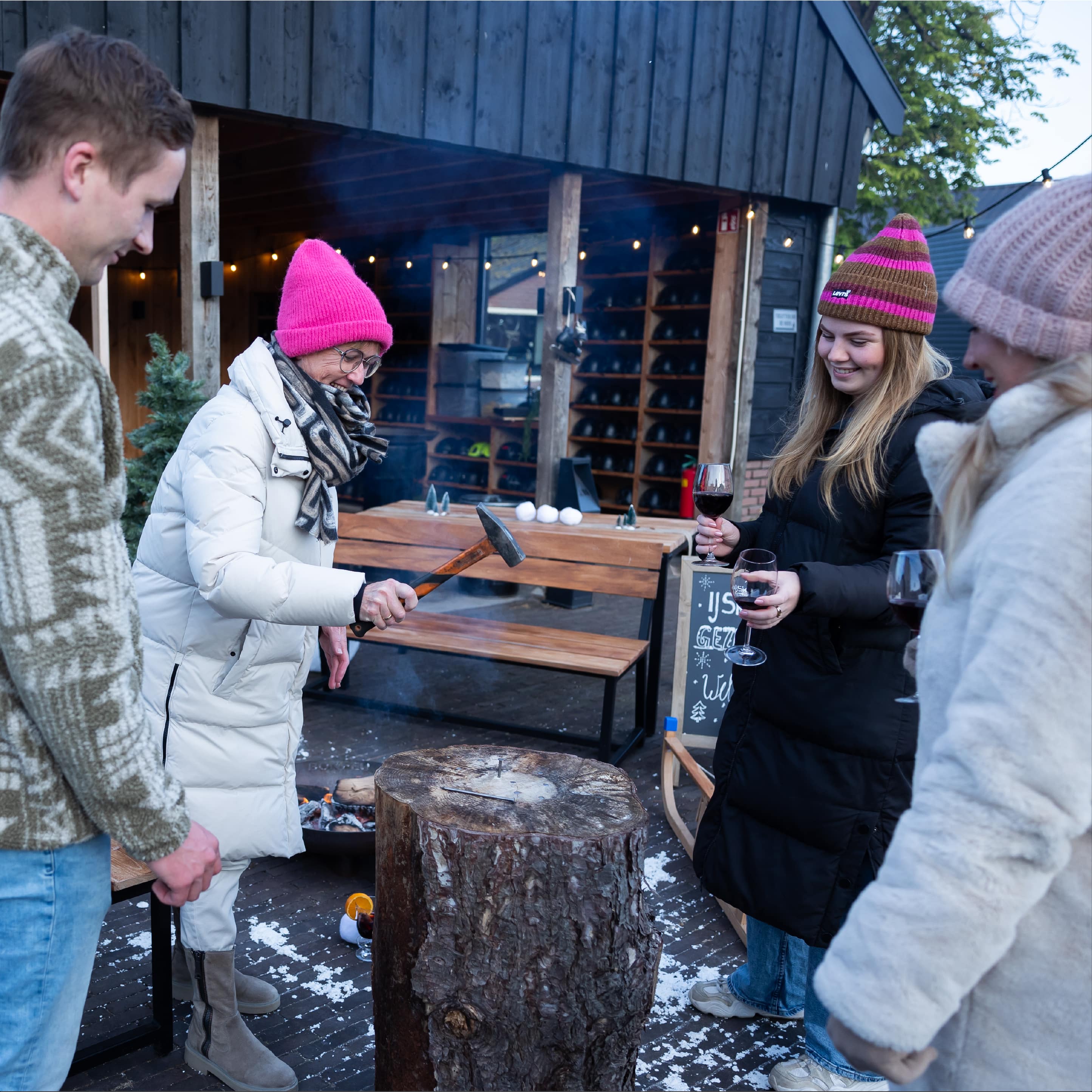 Vier mensen in winterkleding spelen een spel waarbij ze een spijker met een hamer in een boomstronk proberen te slaan, terwijl ze rode wijn drinken.