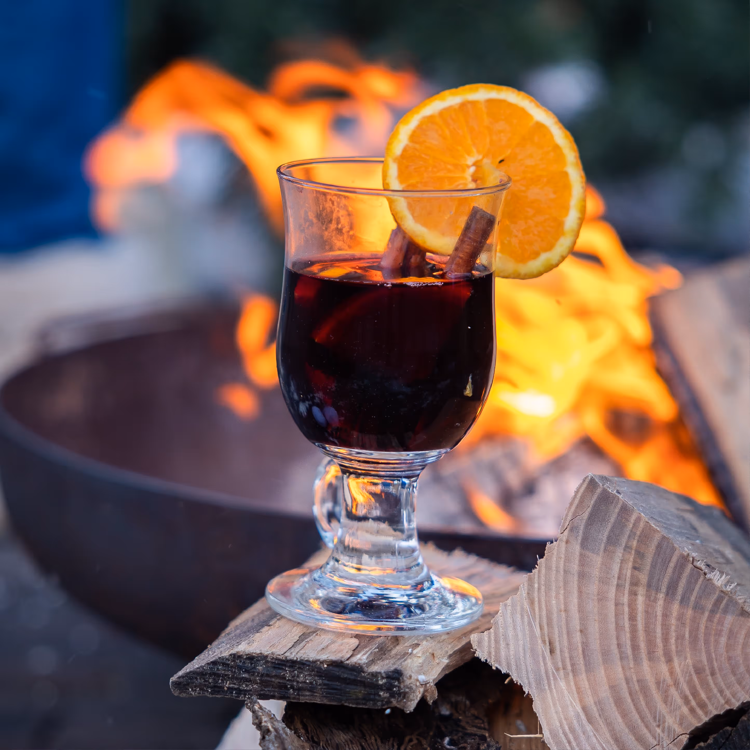 Glas warme glühwein met kaneelstokjes en een schijfje sinaasappel, geplaatst op hout bij een kampvuur.