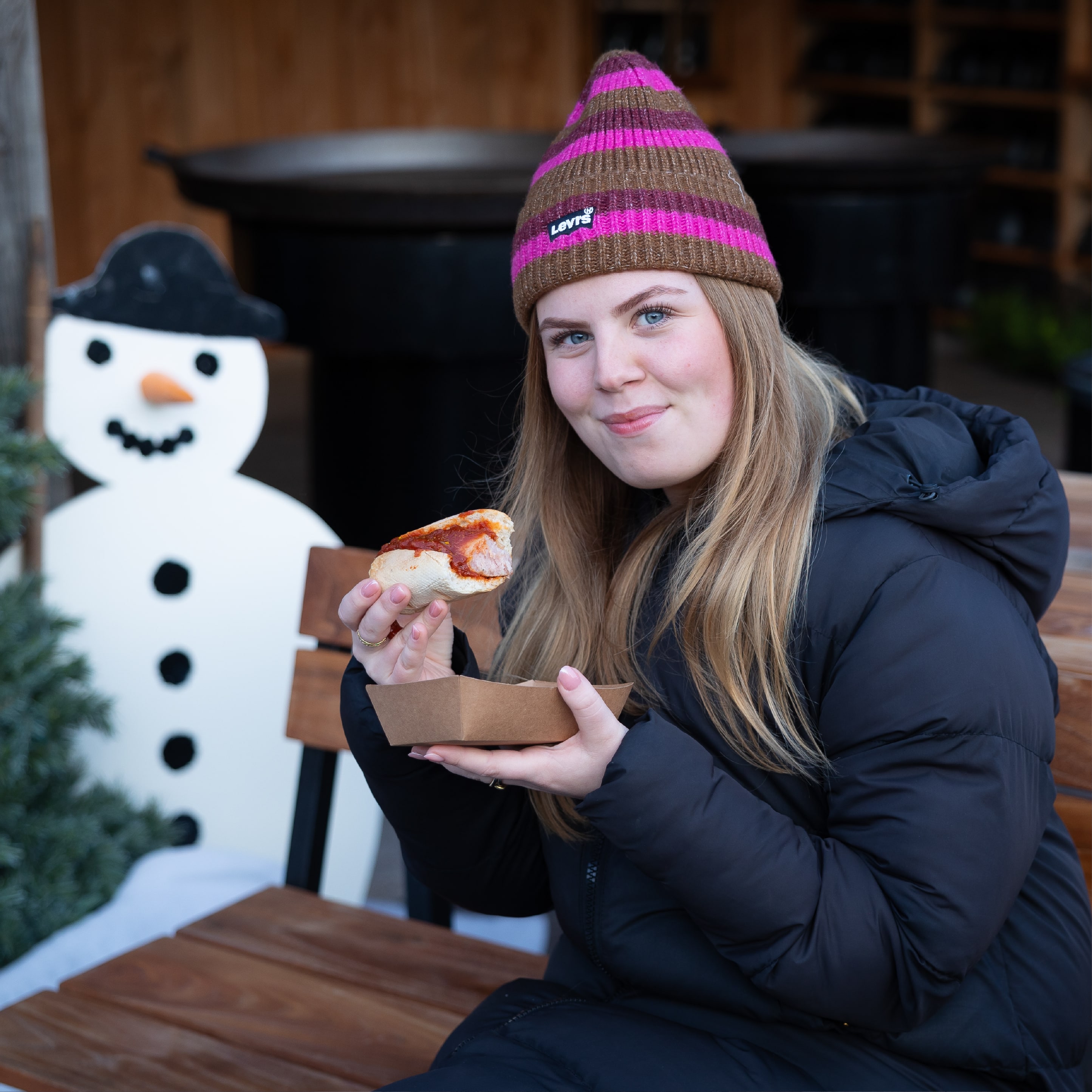 Meisje met een paarse en bruine gestreepte muts eet een stuk pizza terwijl ze buiten zit, met een houten sneeuwpop op de achtergrond.