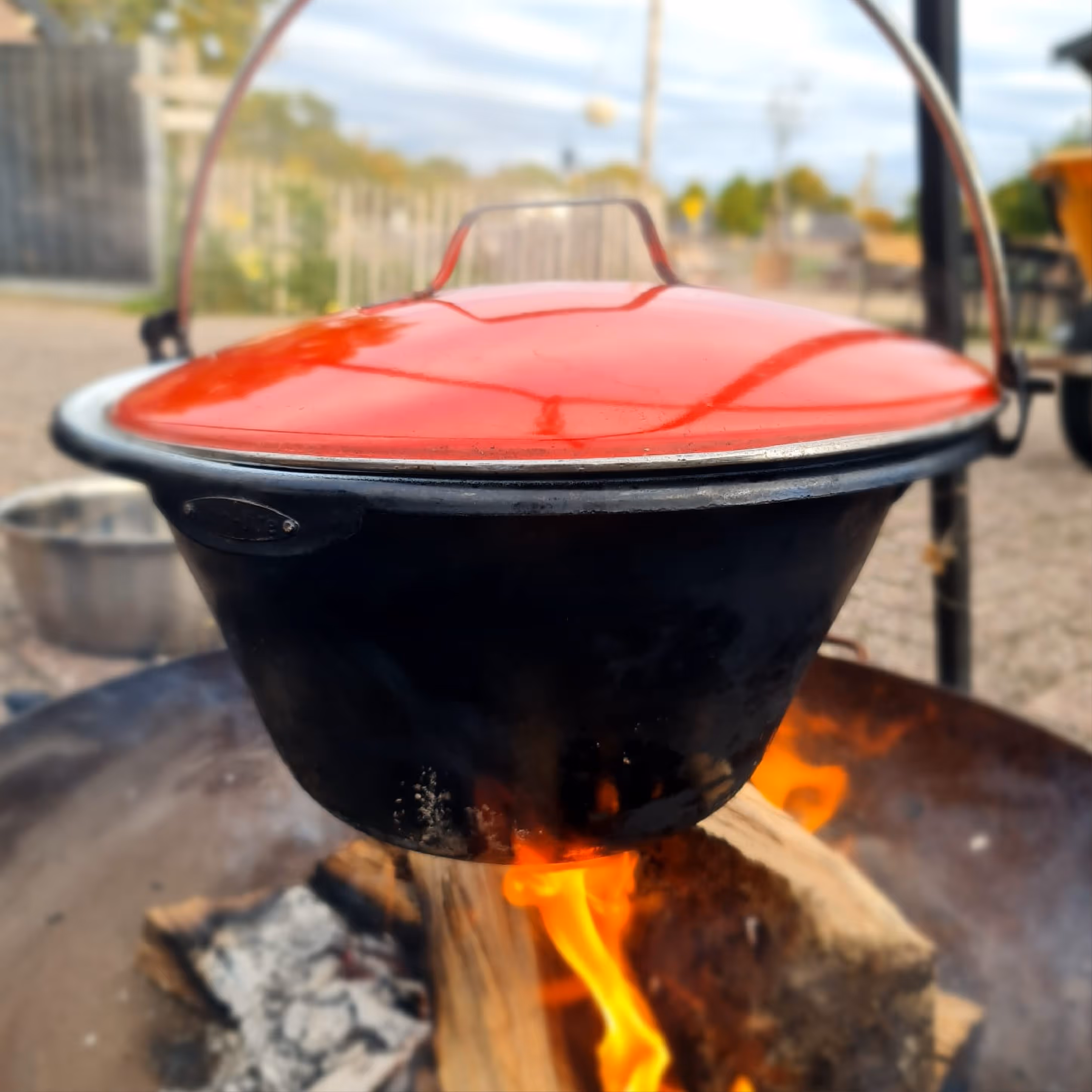 Gietijzeren pot met rode deksel hangt boven een open vuur in een buitenomgeving.