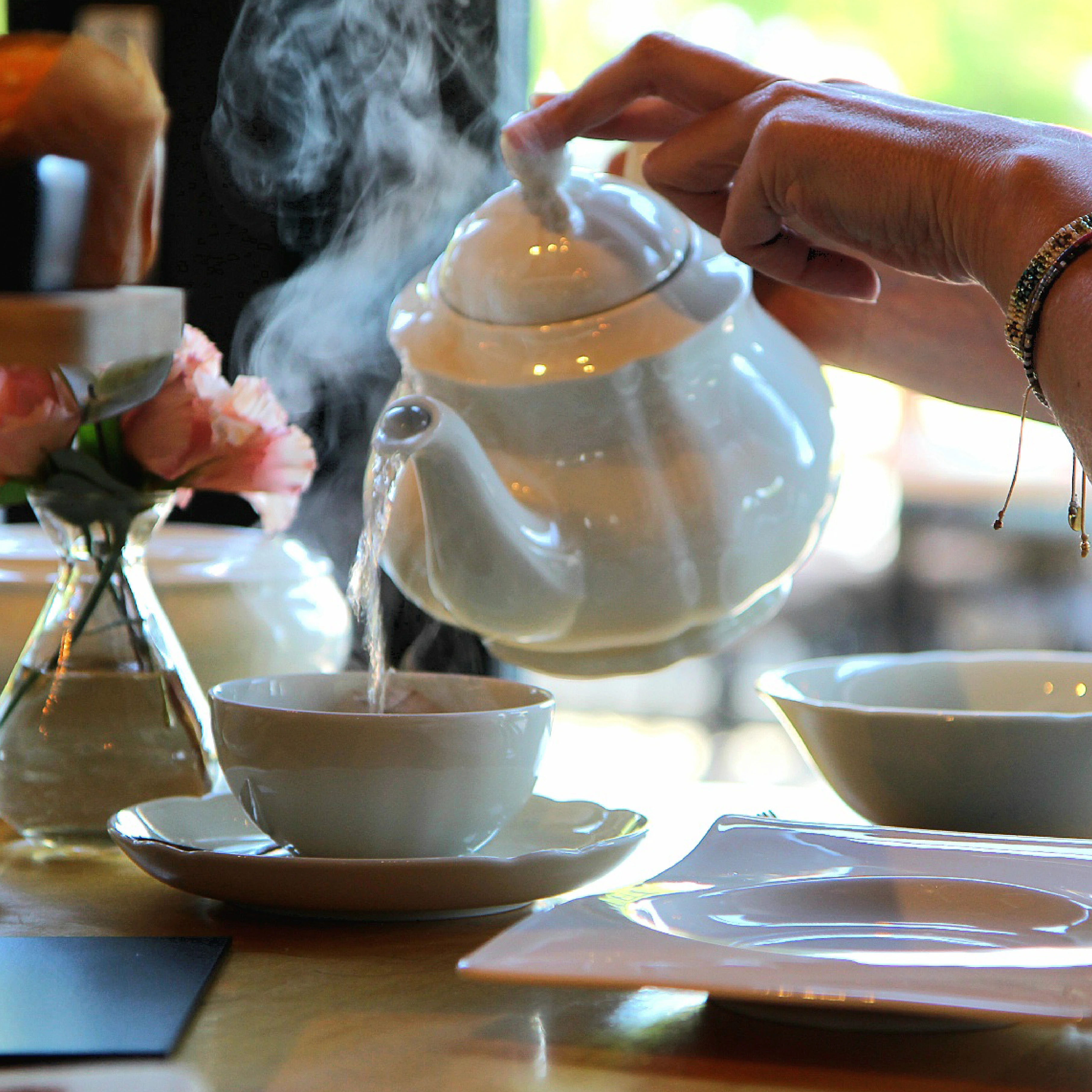 Hand met armbanden schenkt hete thee uit een witte theepot in een kopje op een tafel met bloemen en servies.