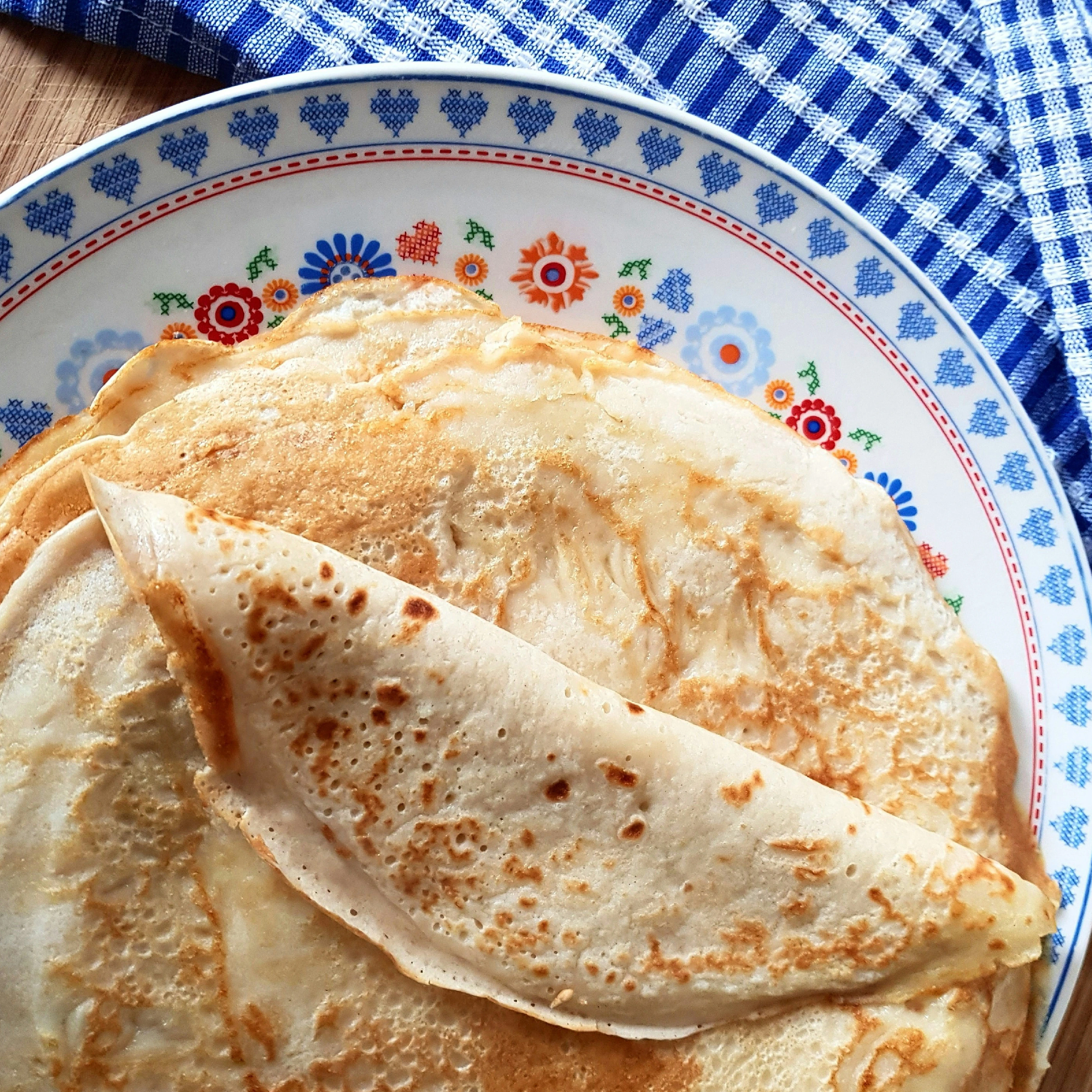 Close-up van een stapel goudbruine pannenkoeken op een bord met blauwe en rode bloemmotieven, op een houten tafel met een blauw geruit keukendoek.