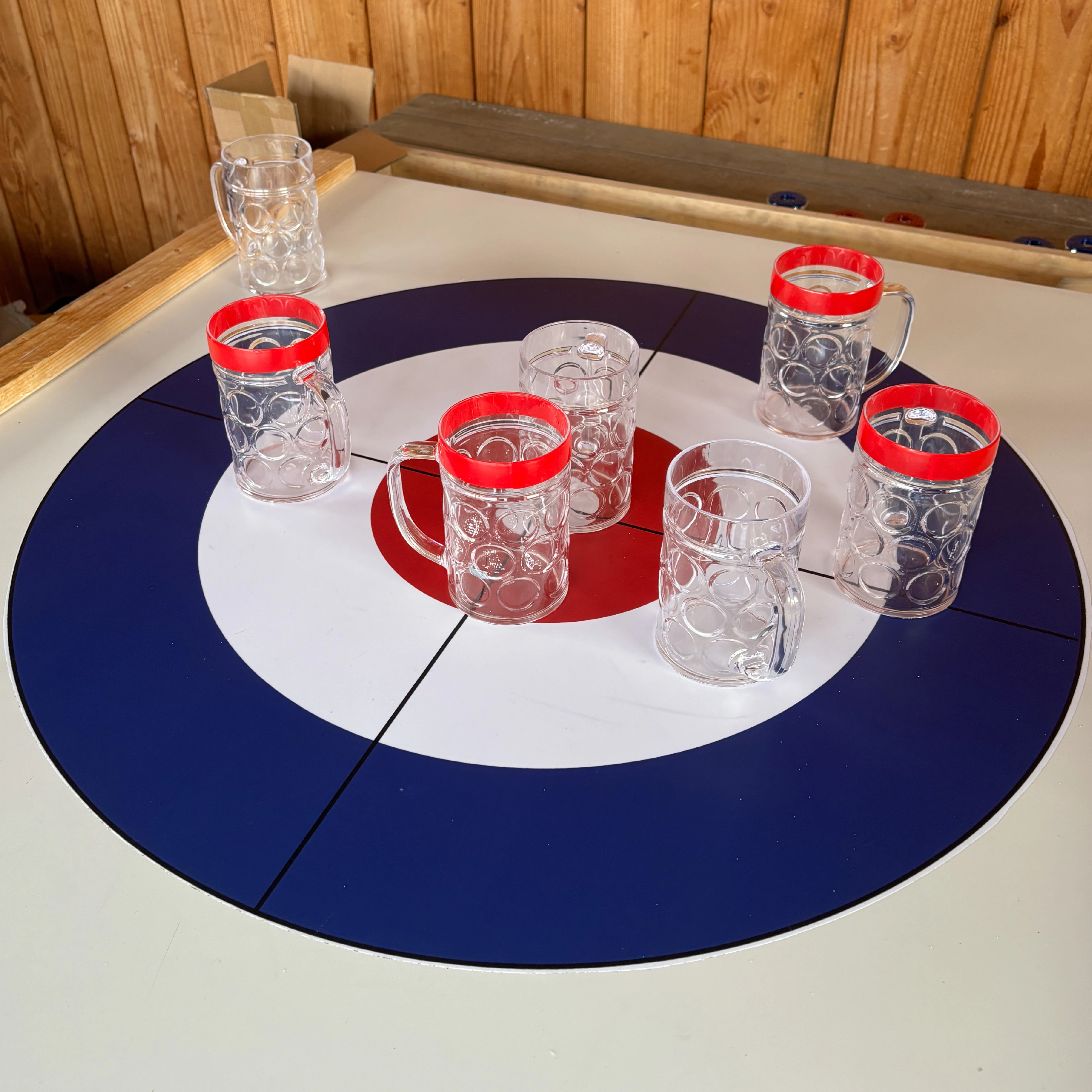 Een curlingbaan met zes glazen biervaten op de cirkel, elk met een rode rand, geplaatst op verschillende posities.