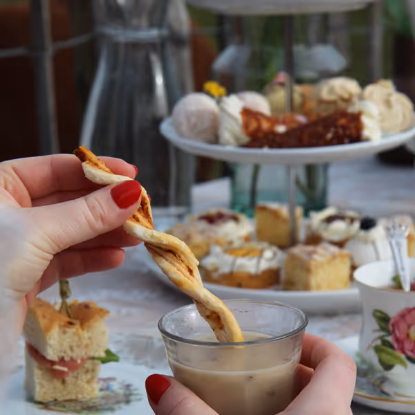 Hand met rode nagellak die een gedraaide snack doopt in een glas melkachtige drank, met een tafel vol hapjes en een theekopje op de achtergrond.