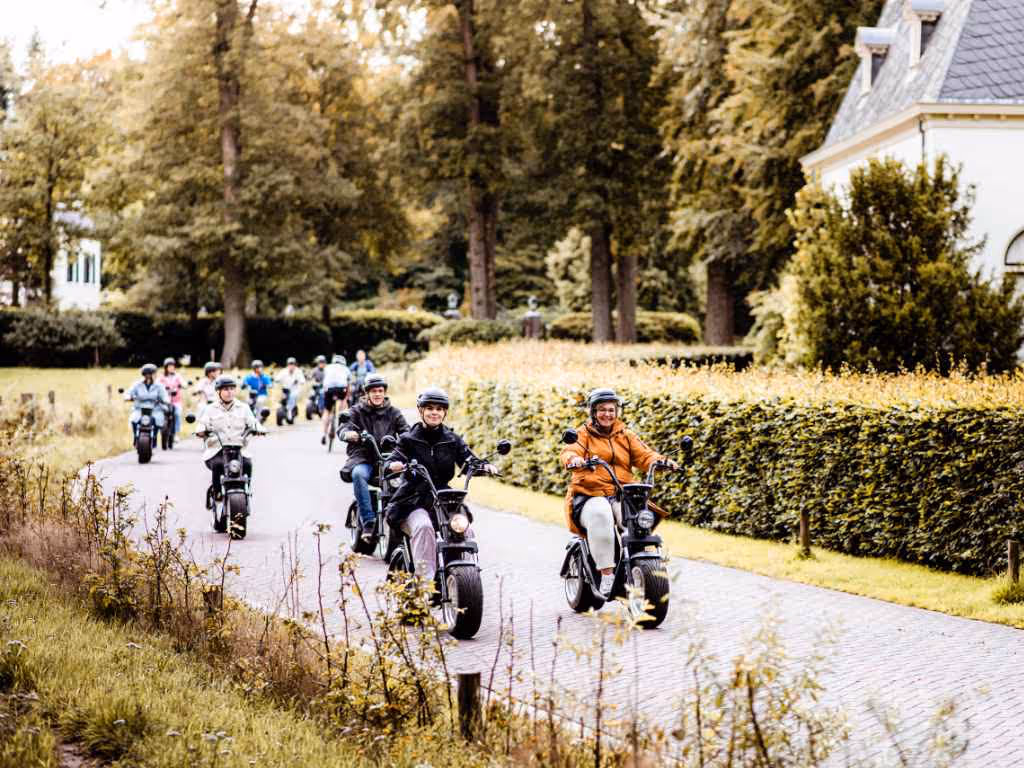 Groep mensen die op elektrische scooters een landweg volgen met groene bomen en struiken aan beide zijden.