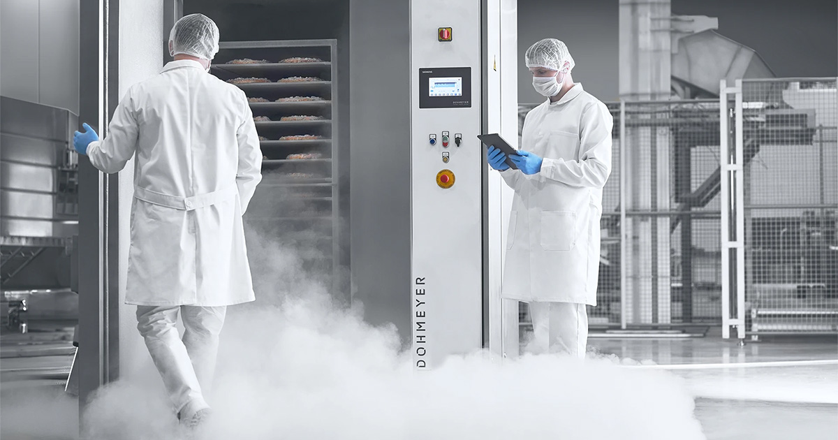 Hygienic food production plant using Dohmeyer cryogenic freezing equipment. Two employees are wearing white lab overalls, blue nitrile gloves, face masks and hair nets, suggesting a high level hygienic production process.