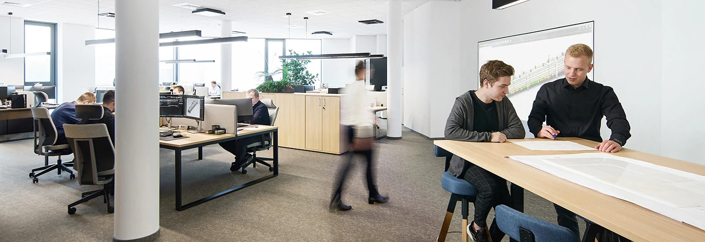 Office space with people sitting at desks, two engineers discussing drawings with model displayed on the back wall