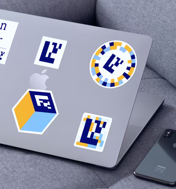 Silver laptop on a grey couch with multiple colorful geometric stickers and an adjacent black smartphone.