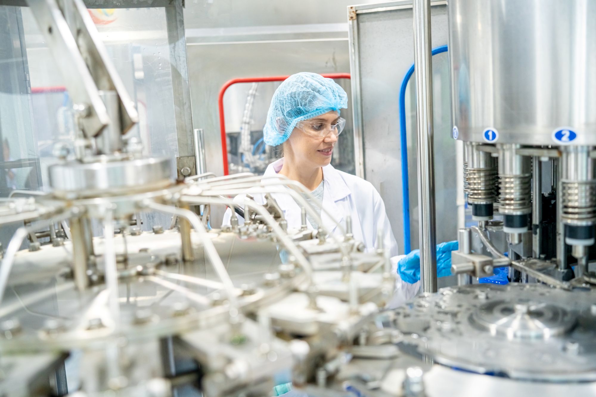 Kvindelig laboratorietekniker iført hårnet, beskyttelsesbriller og handsker betjener maskiner i et sterilt laboratoriemiljø.