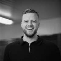 Black and white portrait of a smiling man with short hair and a beard wearing a dark zip-up shirt.