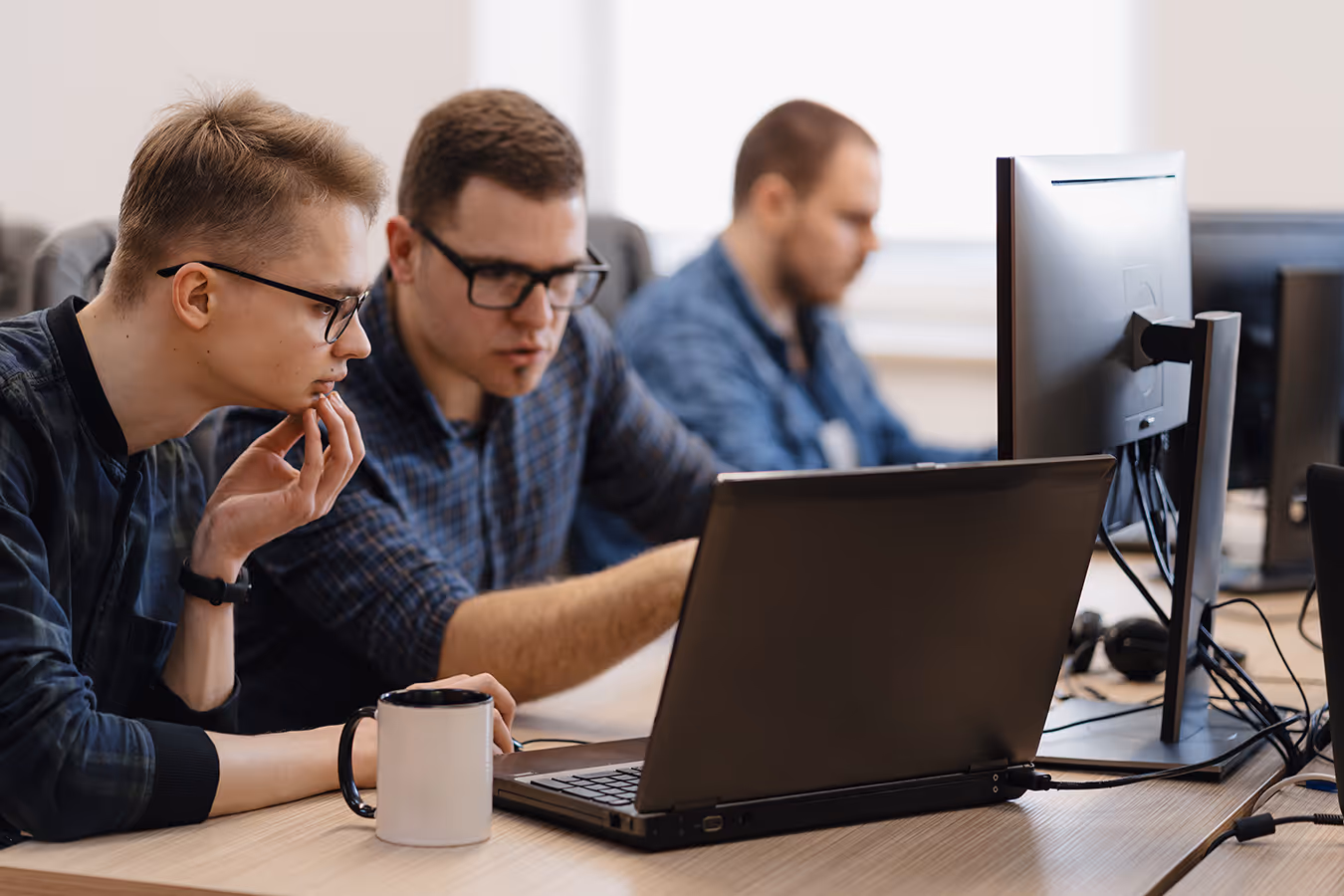 Two men wearing glasses working together on a laptop in an office with another man working on a desktop.