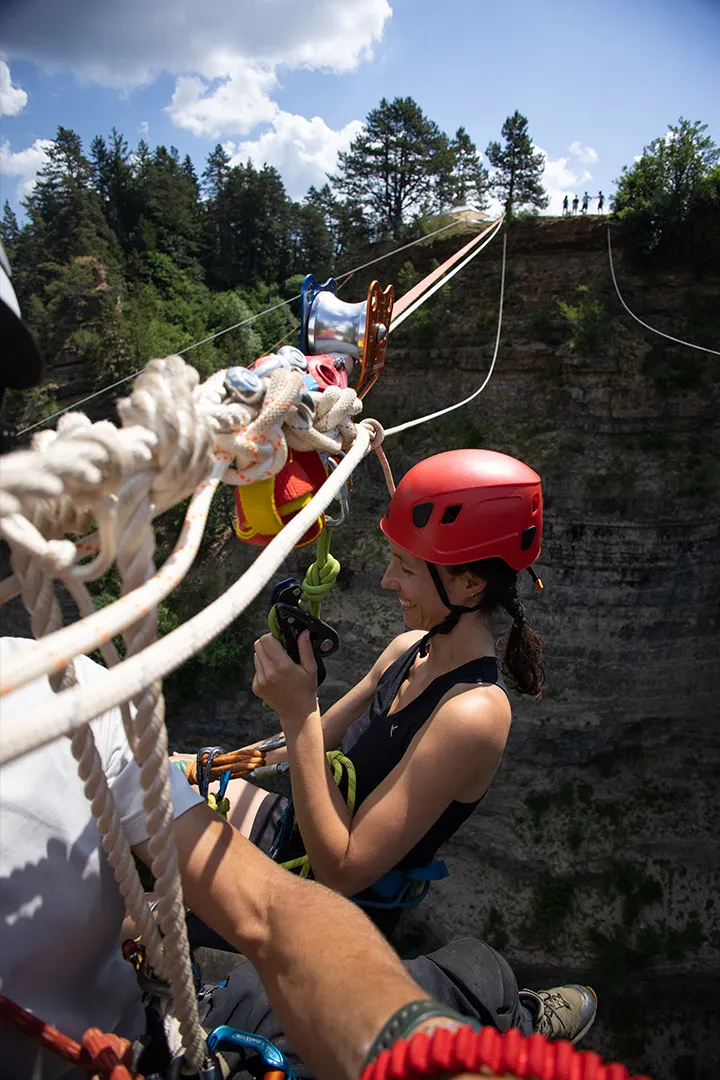 Climber in a red helmet preparing for a rope swing over a rocky cliff with trees above.