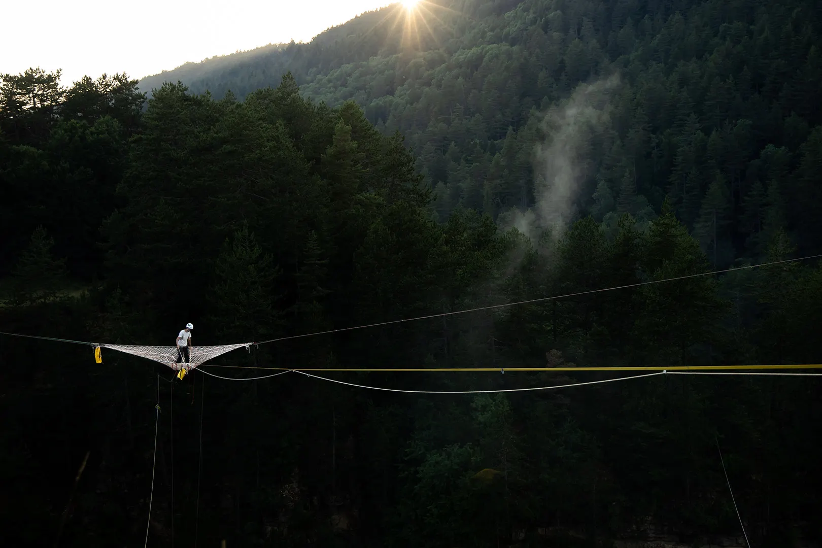 Person in helmet standing on a safety net suspended by ropes over a forested valley at sunset.
