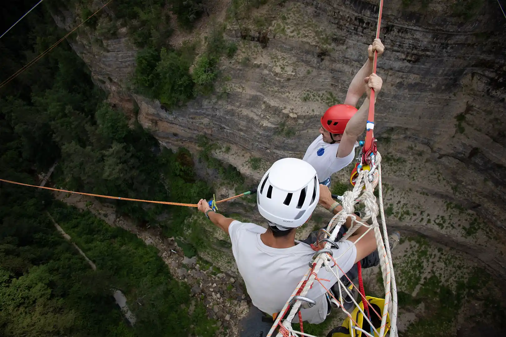 Two people headfirst bungee jumping or pendulum swinging with cliffs and forest below.