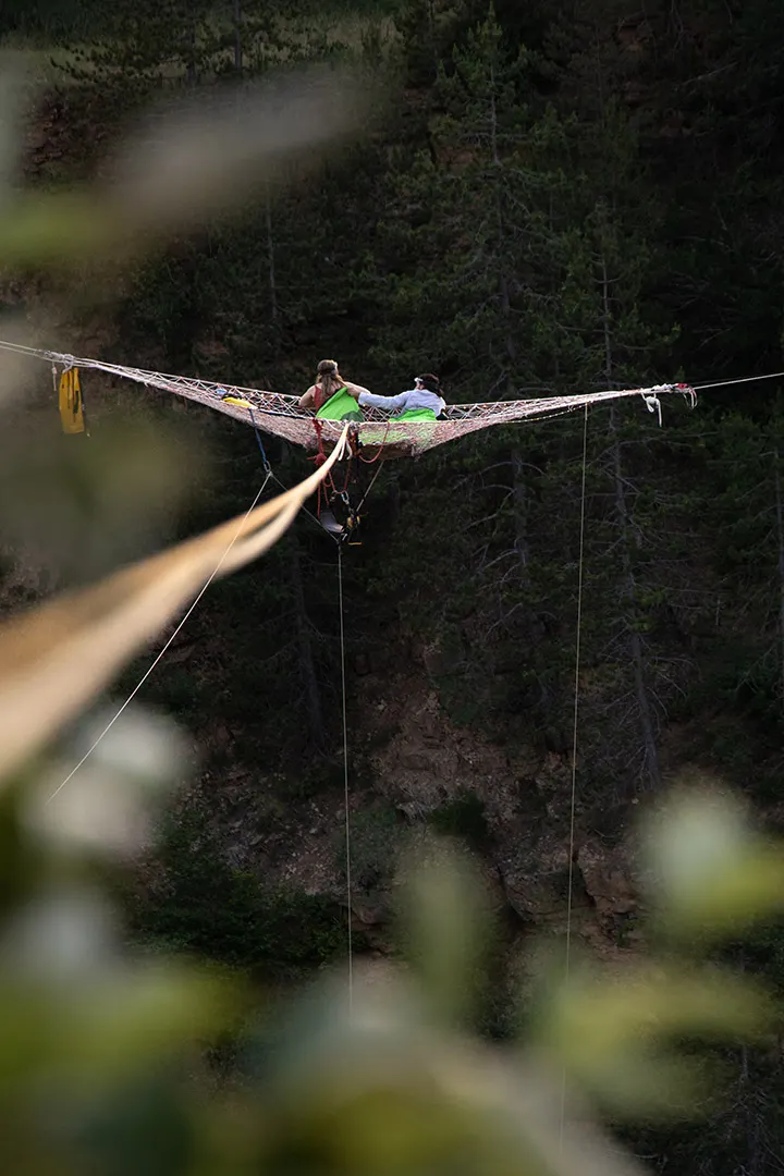 Two people sitting on a large net suspended high above a forested rocky cliff.