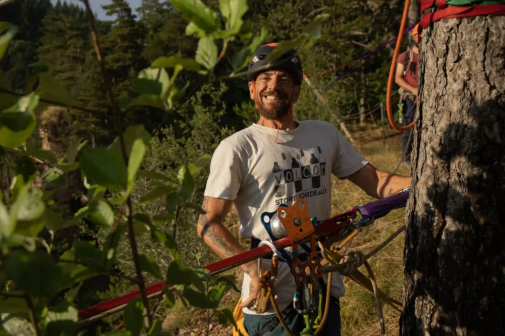 Smiling man in climbing gear holding ropes next to a tree in a forest setting.
