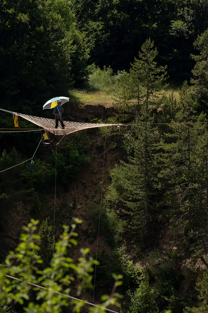 Person walking on a rope net bridge holding a colorful umbrella above a forested canyon.
