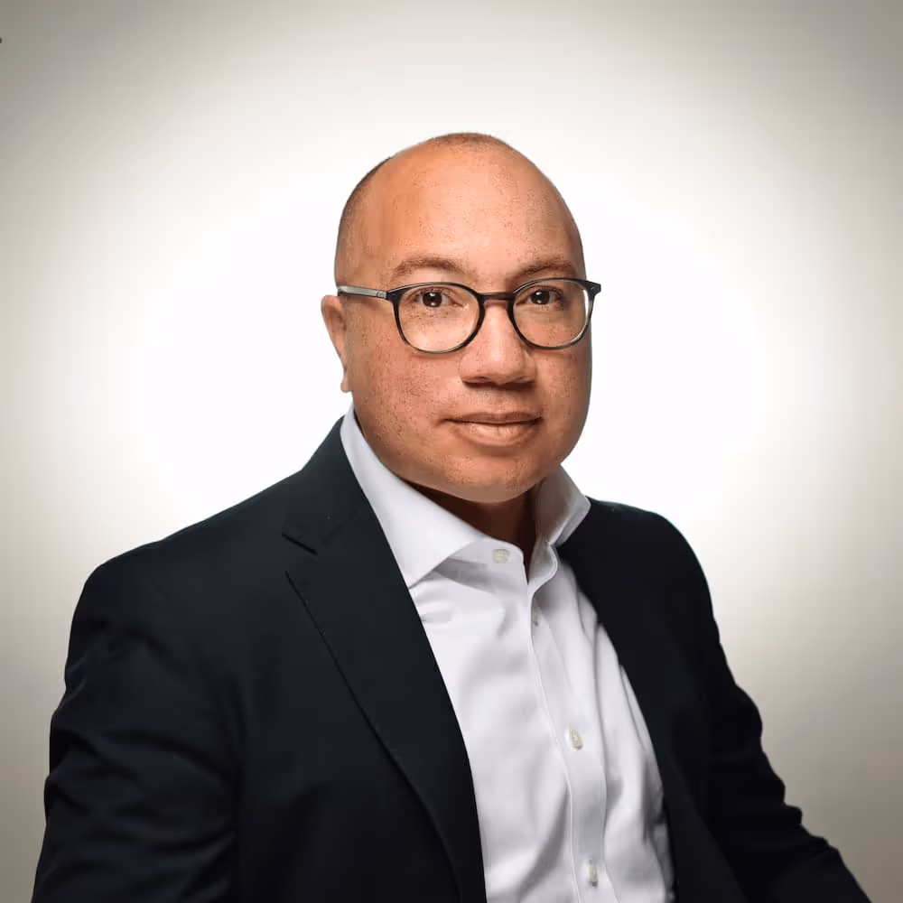 A bald man wearing black-rimmed glasses and a professional outfit consisting of a dark navy blazer over a white collared shirt. He has a slight smile and is photographed against a neutral grey background in a business headshot style