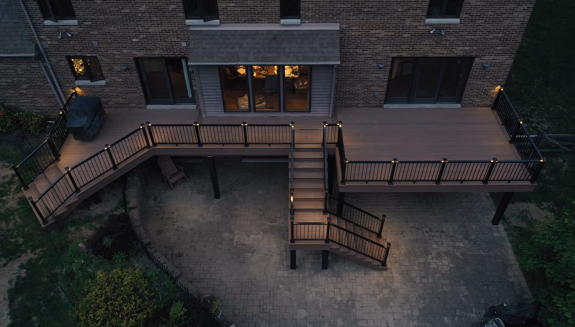 Elevated backyard deck with black railings, stairs, and built-in lights attached to a brick house at dusk.