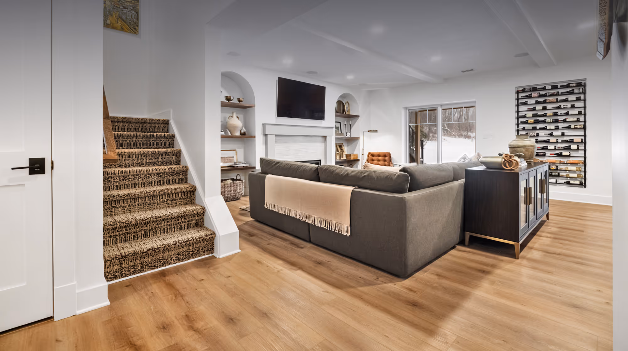 Modern basement living area with gray sectional sofa, wall-mounted TV above a white fireplace, patterned carpeted stairs, wooden floor, and wine racks on the wall.