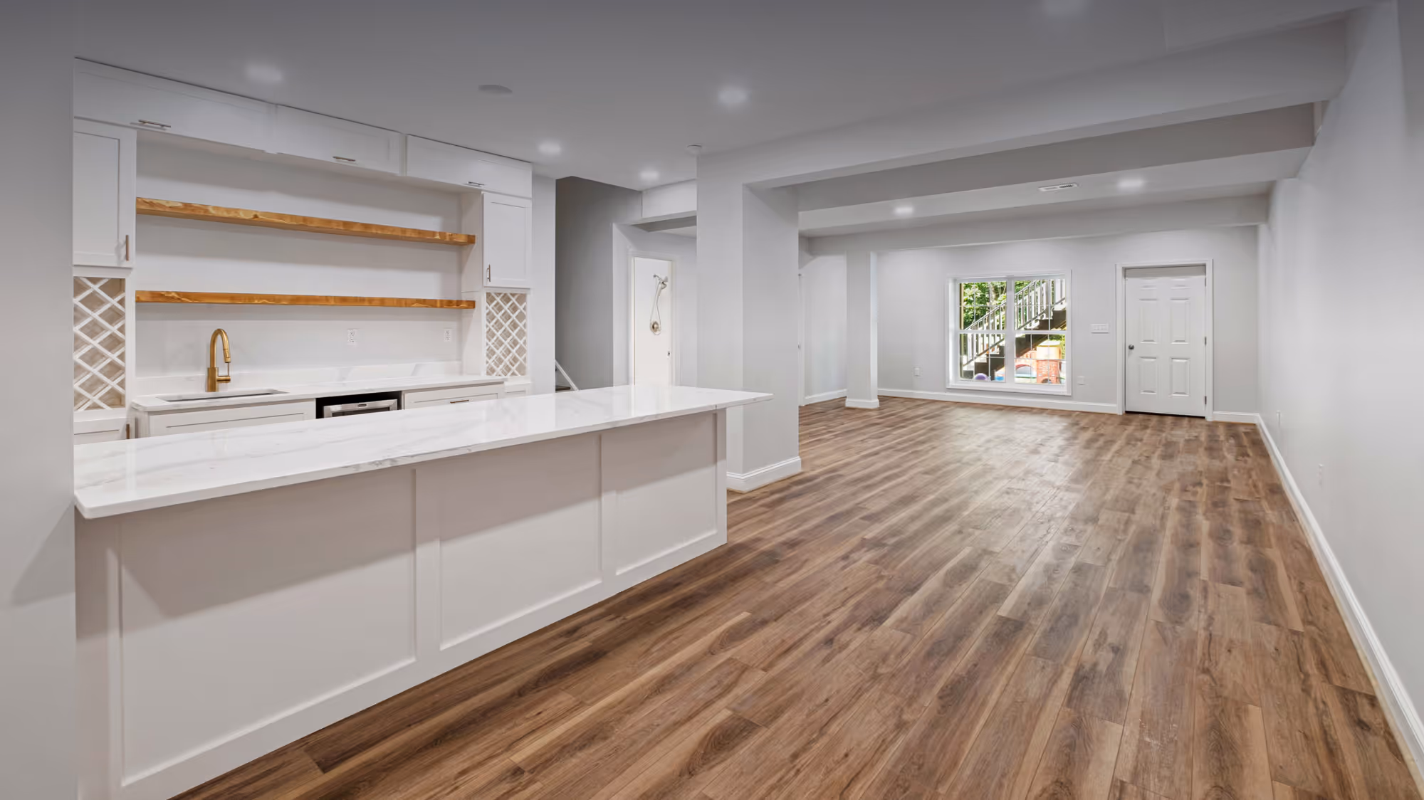 Spacious modern basement with white kitchen island, wooden shelves, and brown wood flooring.