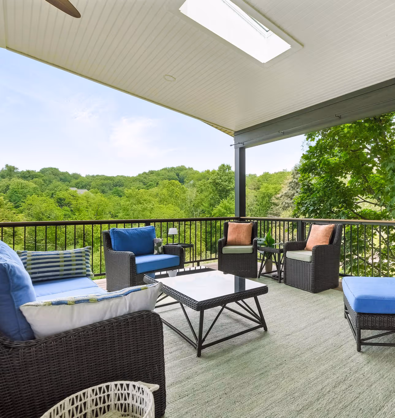 Covered outdoor patio with wicker seating, blue and pink cushions, glass-top coffee table, and green trees in the background.