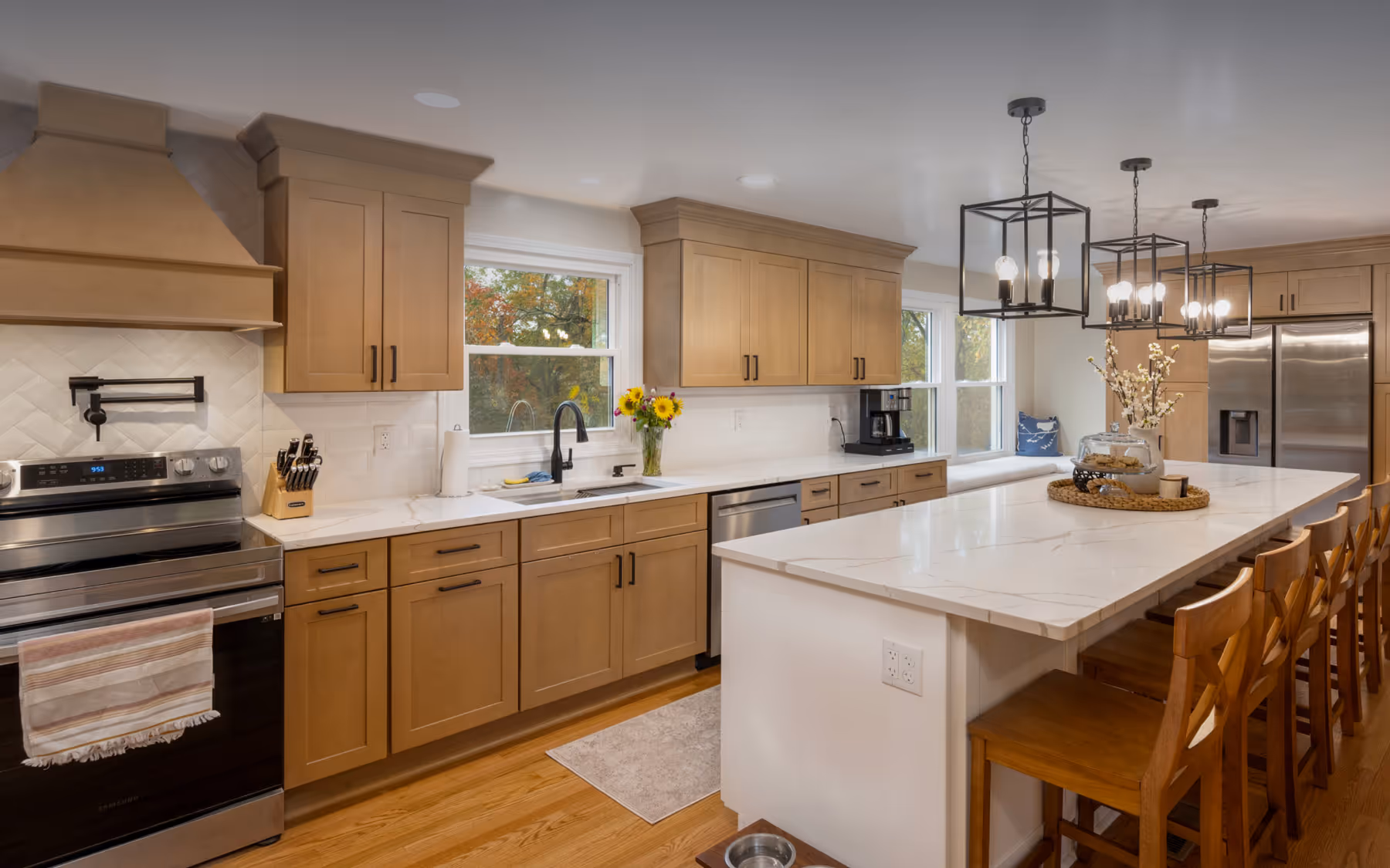 Modern kitchen with wooden cabinets, stainless steel appliances, white marble countertops, pendant lighting, and a long island with wooden bar stools.